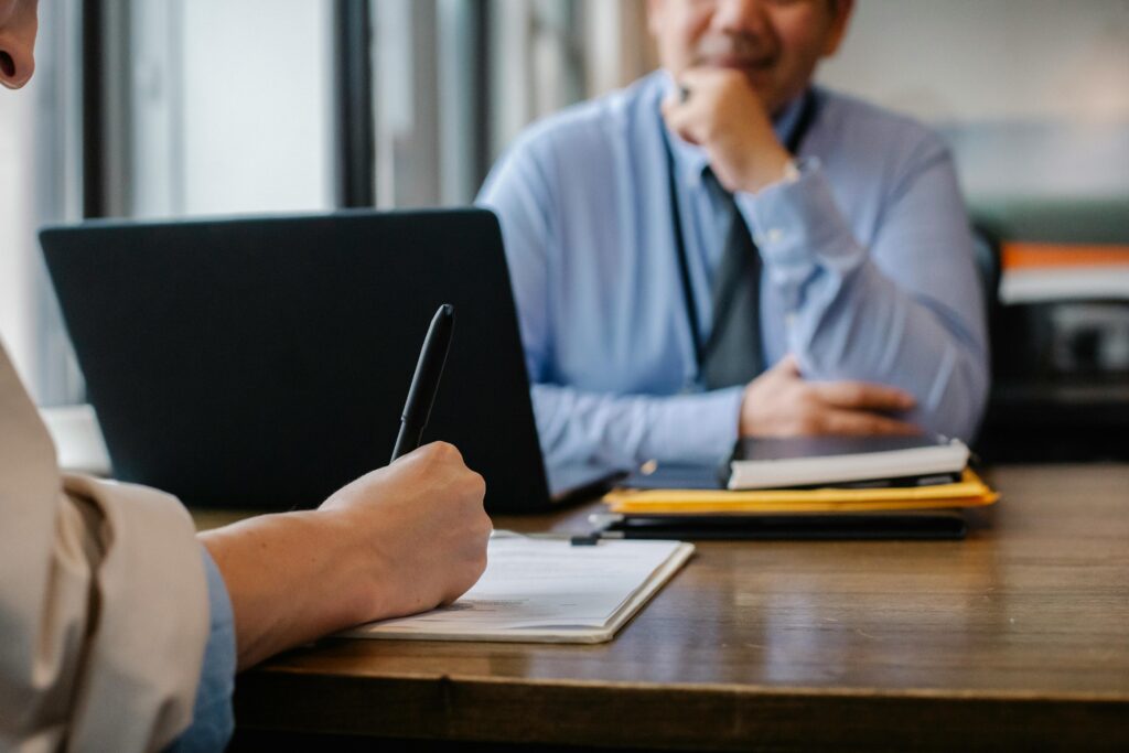 Two people sit either side of a desk in an office. The person cloest to the camera is wearing a white shirt and making notes on a piece of paper. The person opposite them is wearing a blue shirt. Their faces are not visible.