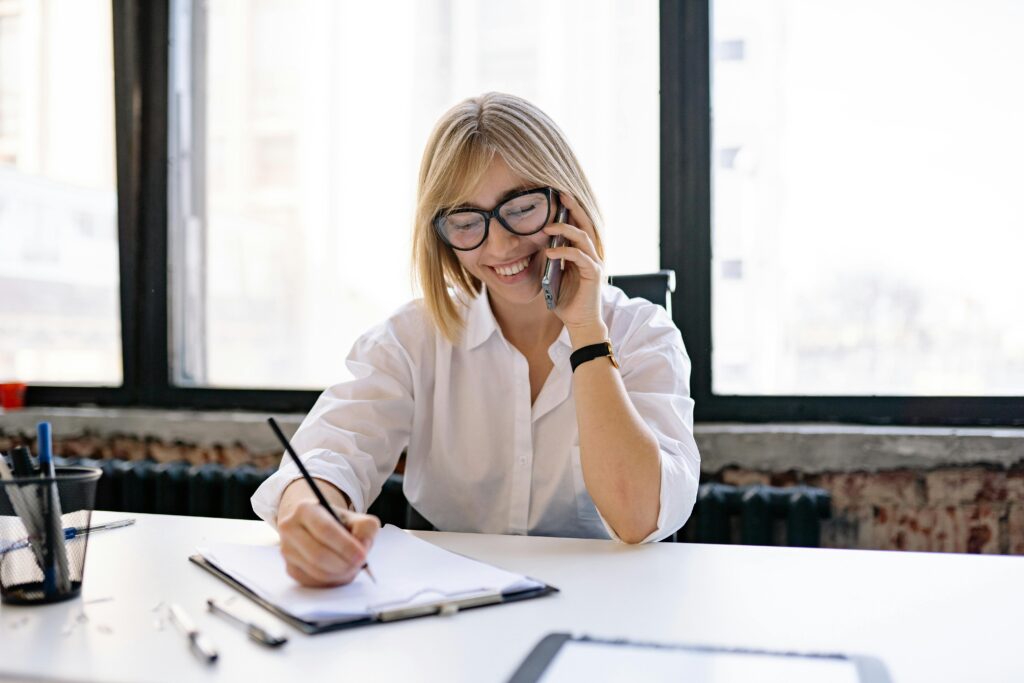 A woman with blonde hair, wearing black glasses, sits behind a desk. She is smiling and speaking to someone on her mobile phone while taking notes on a clipboard.